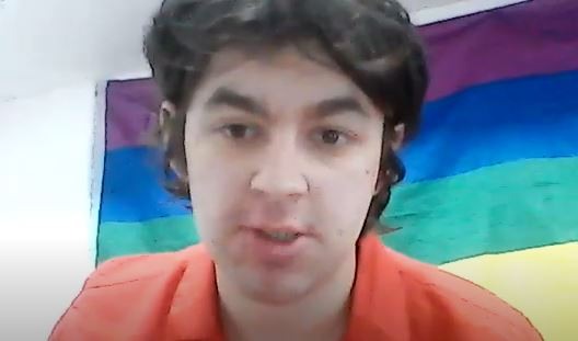 A screenshot of Kieron talking to camera, sitting in front of a Pride flag